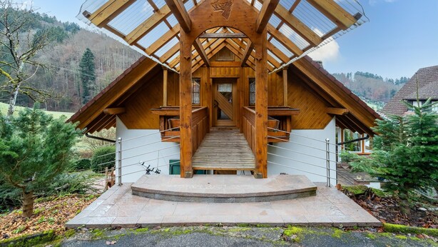 Una cabaña de madera con una escalera que lleva a un porche cubierto, rodeada de árboles y un paisaje montañoso.