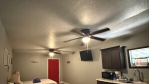Une chambre d’hôtel avec deux lits, une kitchenette, un ventilateur de plafond et un téléviseur fixé au mur.