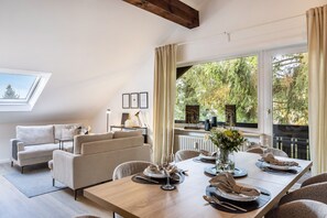 A dining area with a wooden table set for four, a sofa, and a view of trees through large windows.