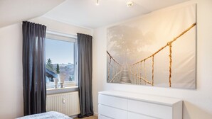 Een slaapkamer met een bed, een witte kapstok, een groot raam met gordijnen en een schilderij van een touwbrug.