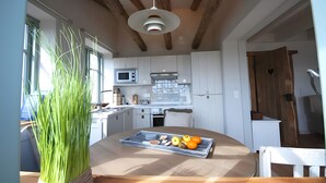 A modern kitchen with white cabinets, a wooden ceiling, and a dining table with a tray of fruits and vegetables. - Port on the Fehmarnsund – Sea View and Beach at Your Doorstep (Fehmarn)