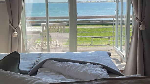 A bedroom with a large window overlooking a beach and a deck. - Port on the Fehmarnsund – Sea View and Beach at Your Doorstep (Fehmarn)