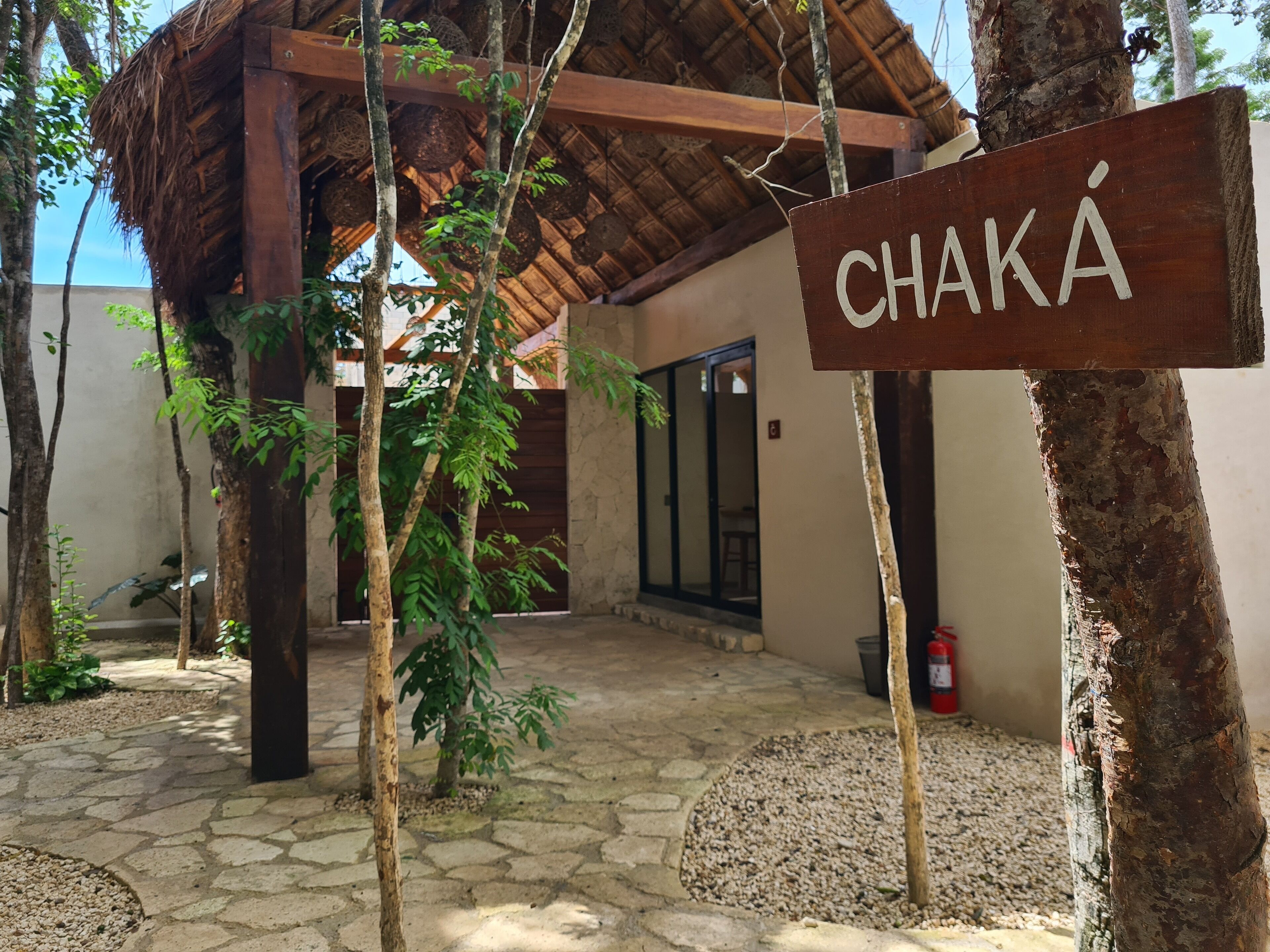 Un letrero de madera con el nombre "CHAKA" en un entorno rústico, con una estructura de techo de paja, suelo de piedra y follaje circundante.