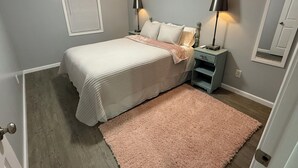 A bedroom with a bed, bedside lamps, a nightstand, a wall-mounted air conditioner, a window with blinds, and a door.
