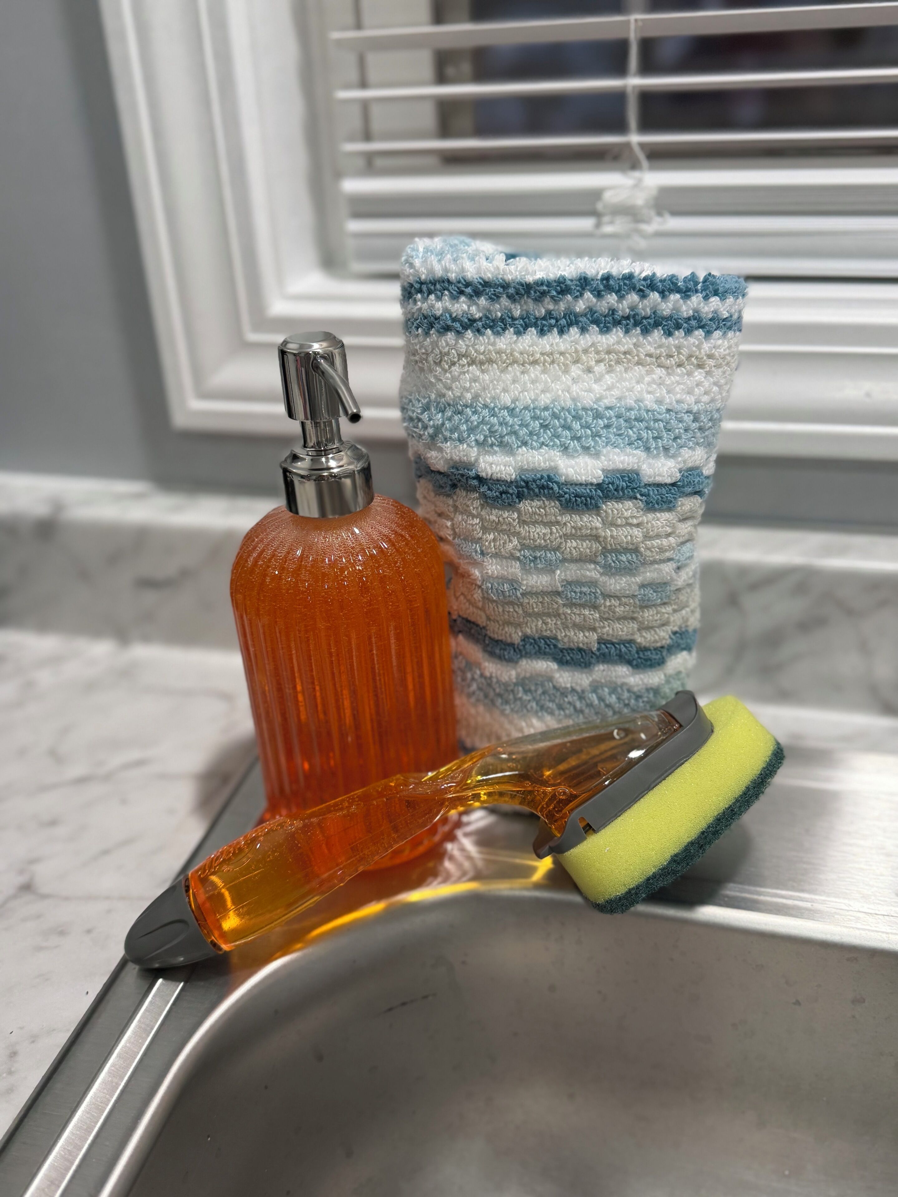 A kitchen sink with a bottle of orange liquid, a yellow and green sponge, and a blue and white striped towel.