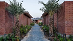 A pathway flanked by brick buildings with palm trees, leading to a covered area. - The Bhagwati Cottage & Villa (Abu Road)