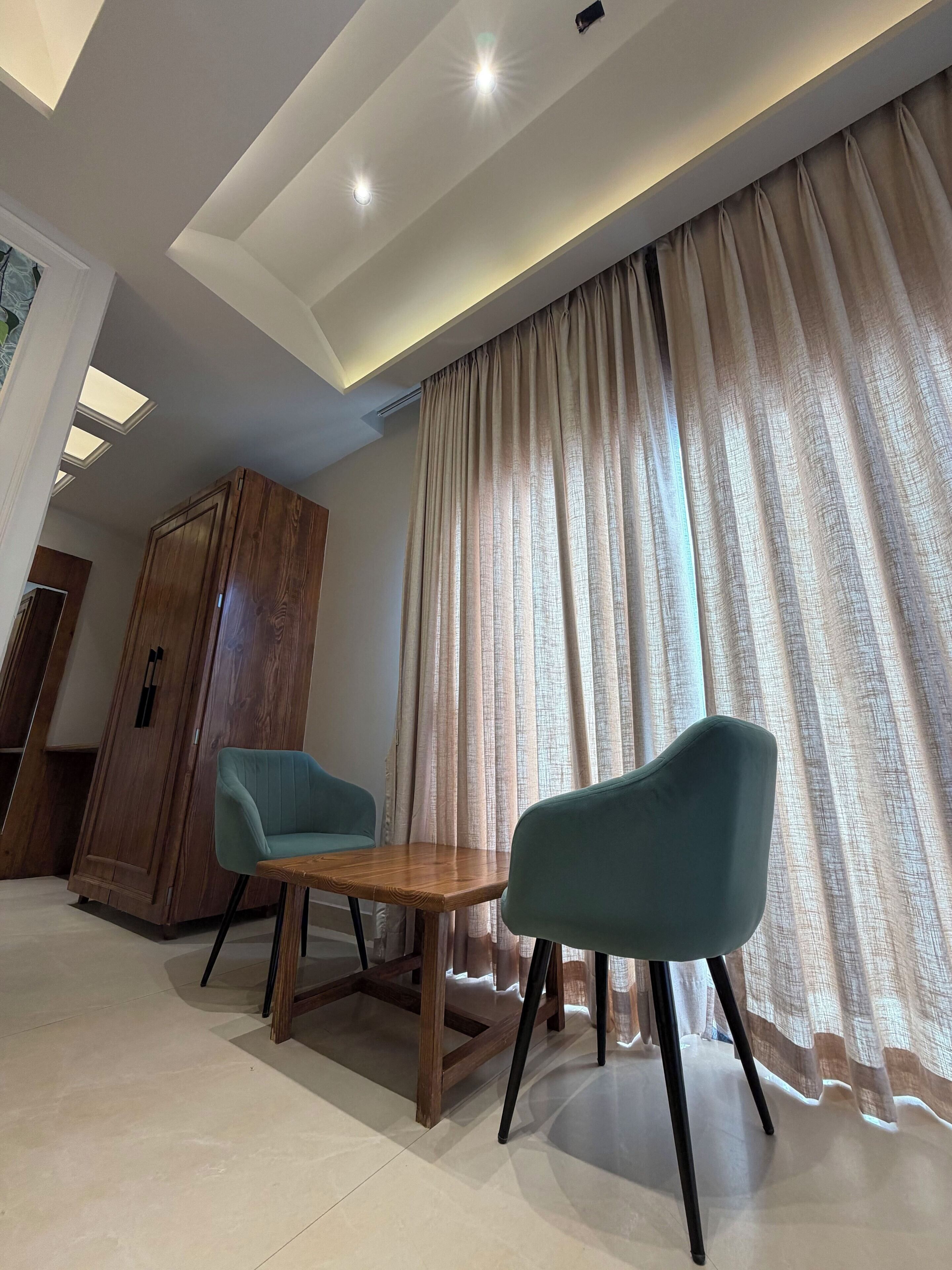 A modern room with a wooden cabinet, two teal chairs, a wooden table, and a large window with curtains.