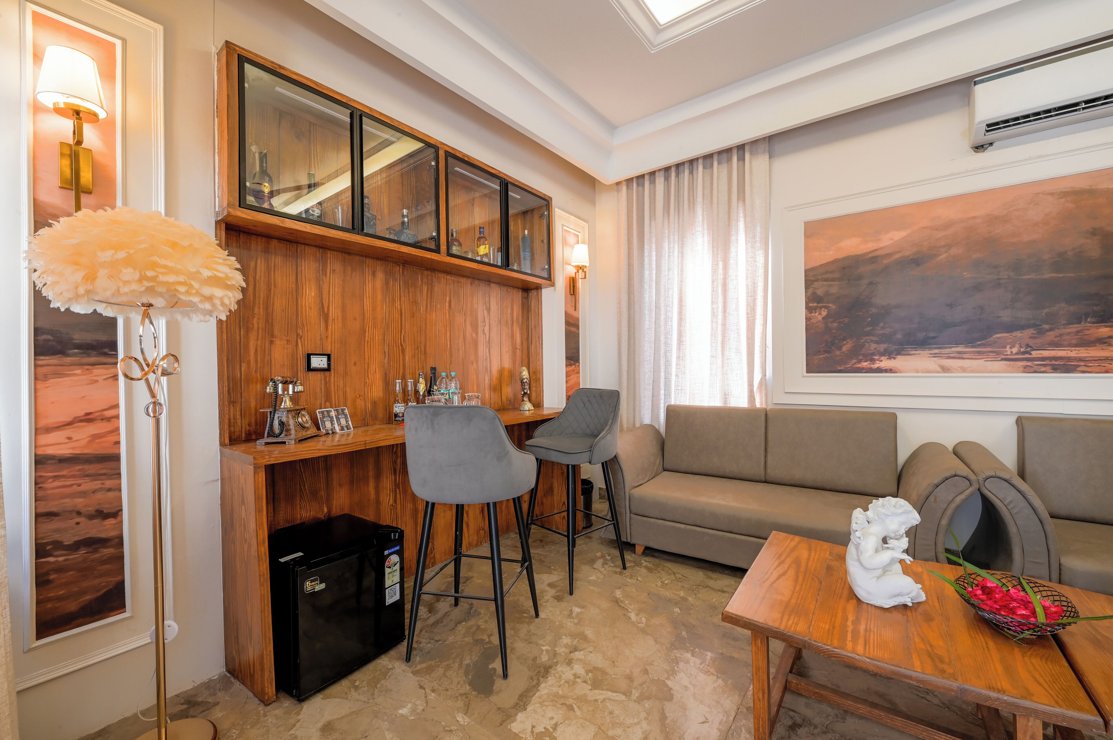 A hotel room with a wooden bar, grey chairs, a sofa, and a table with a decorative item and flowers.
