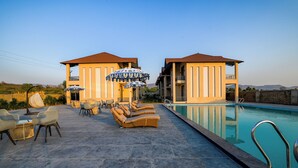 A poolside area with lounge chairs and umbrellas, a pool, and two buildings with balconies. - The Bhagwati Cottage & Villa (Abu Road)
