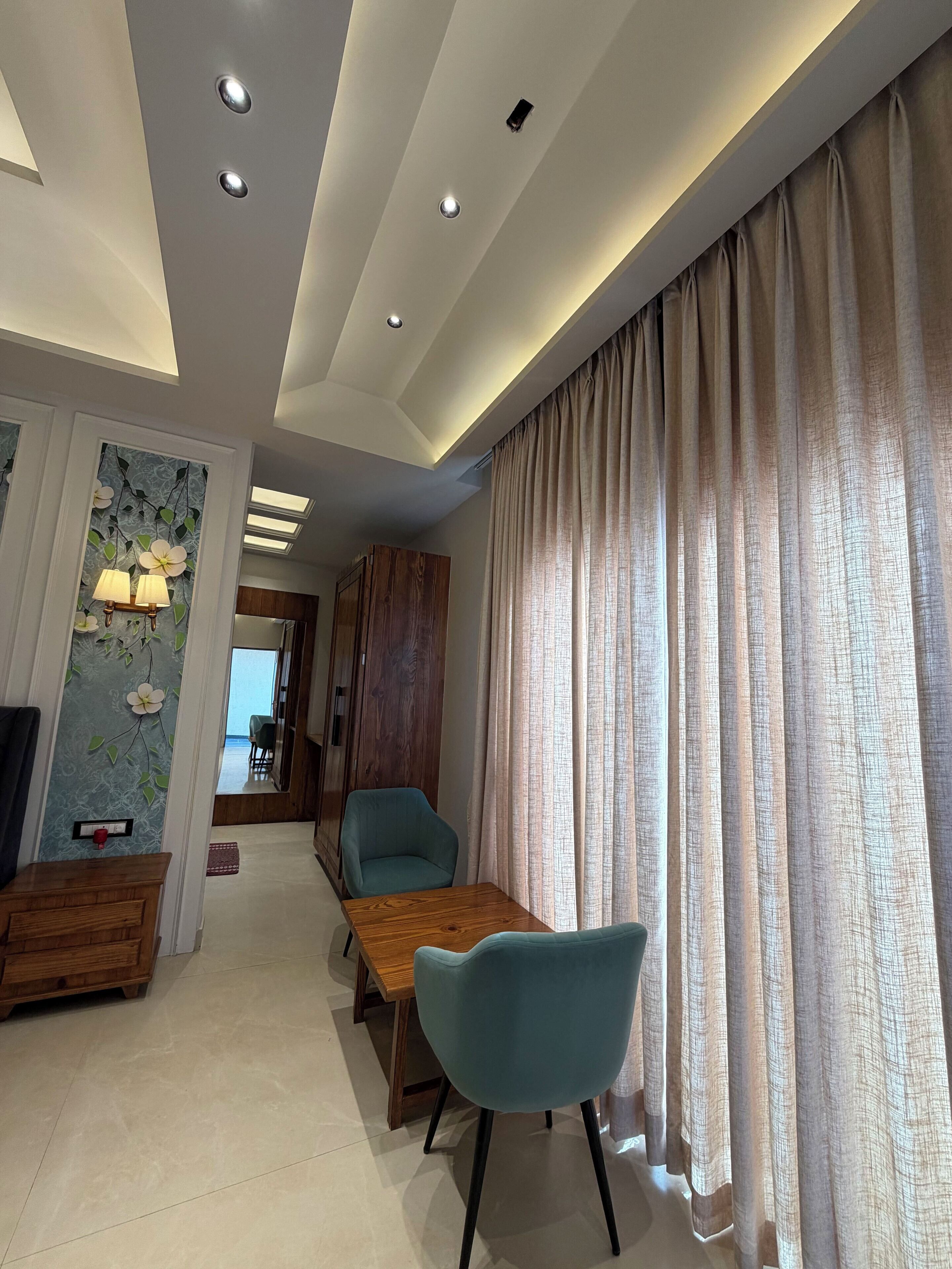 A modern dining area with a wooden table and blue chairs, a ceiling with recessed lighting, and a view of a balcony through a window with curtains.