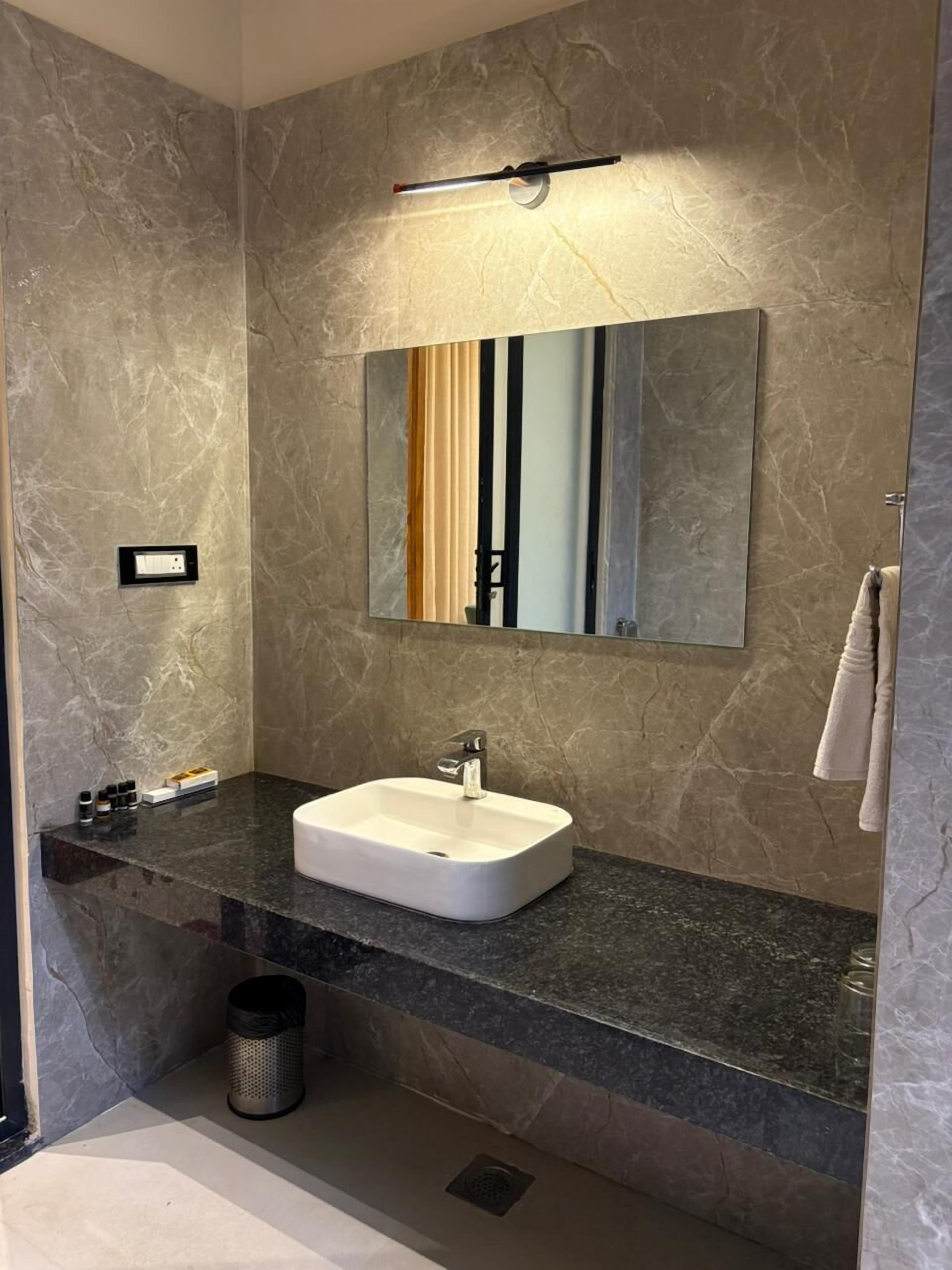 Un baño moderno con pared de mármol, un espejo grande, un lavamanos blanco y un cubo de basura negro.