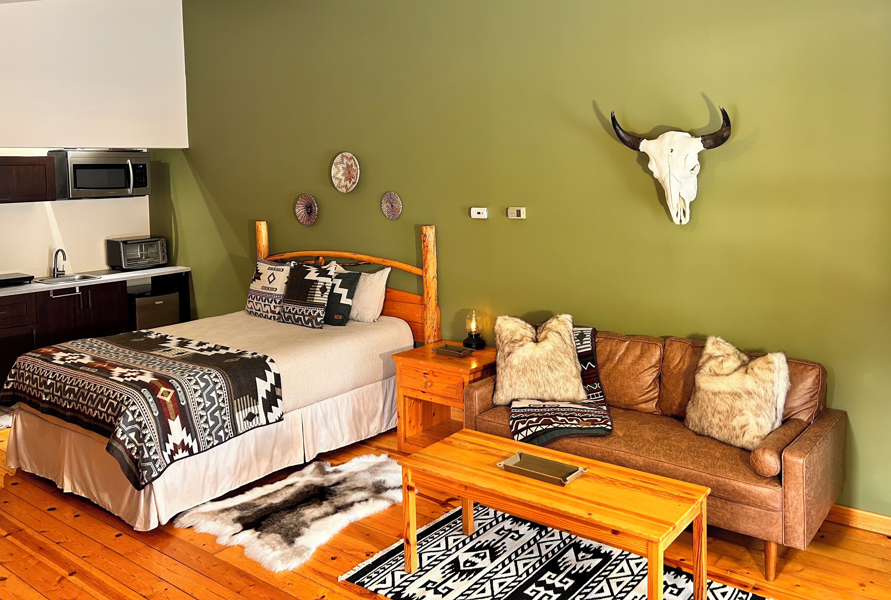 A room with a bed, a sofa, a coffee table, a wall-mounted microwave, a small table with a lamp, and a wall-mounted bull skull.