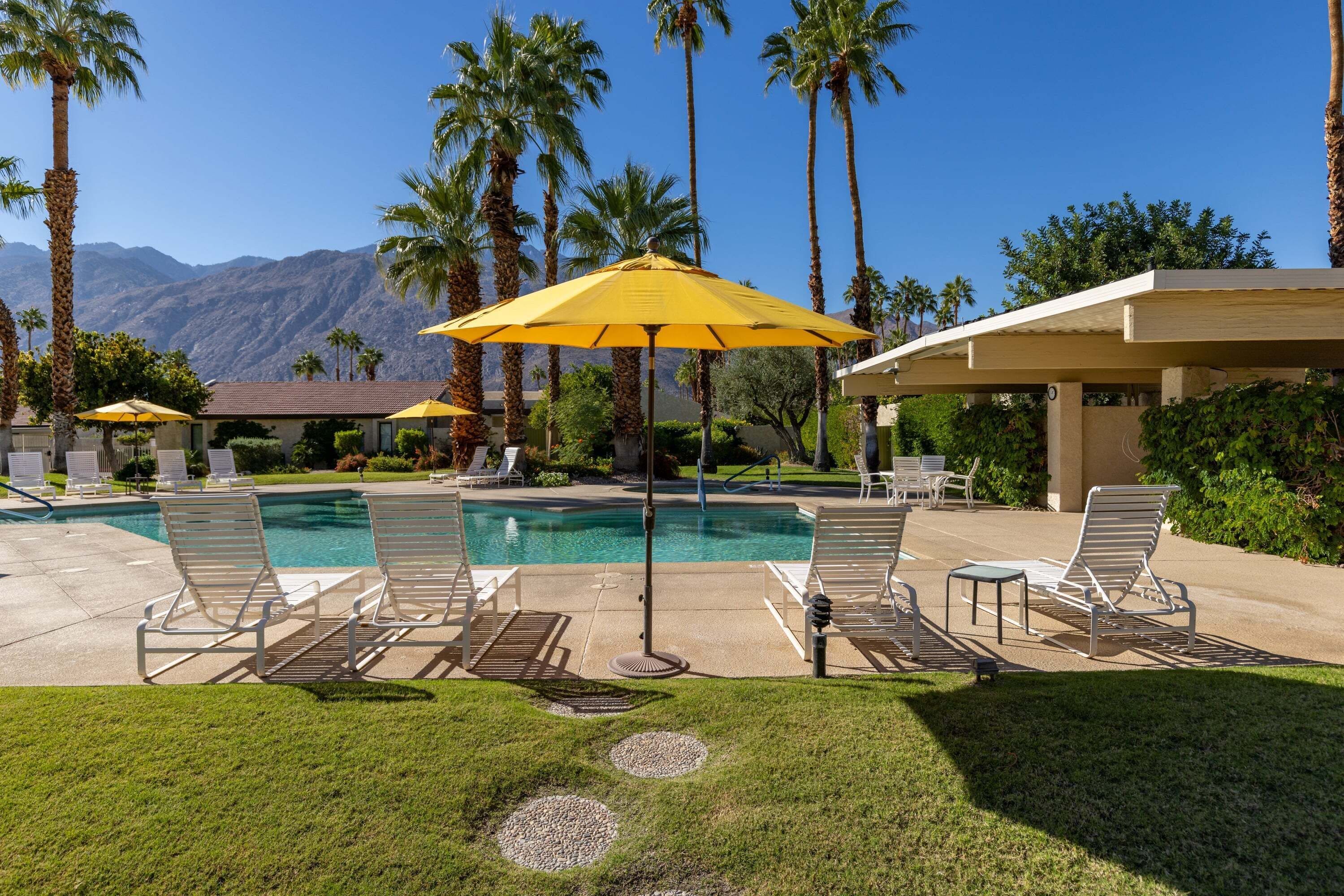 Zona de piscina con sillones de descanso, sombrillas y palmeras.