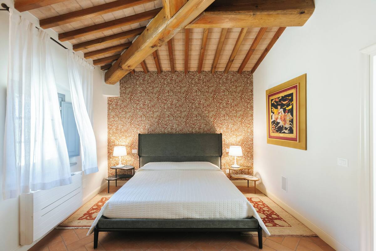 A bedroom with a bed, two bedside tables, a nightstand, a window with curtains, a patterned wall, and a framed picture on the wall.