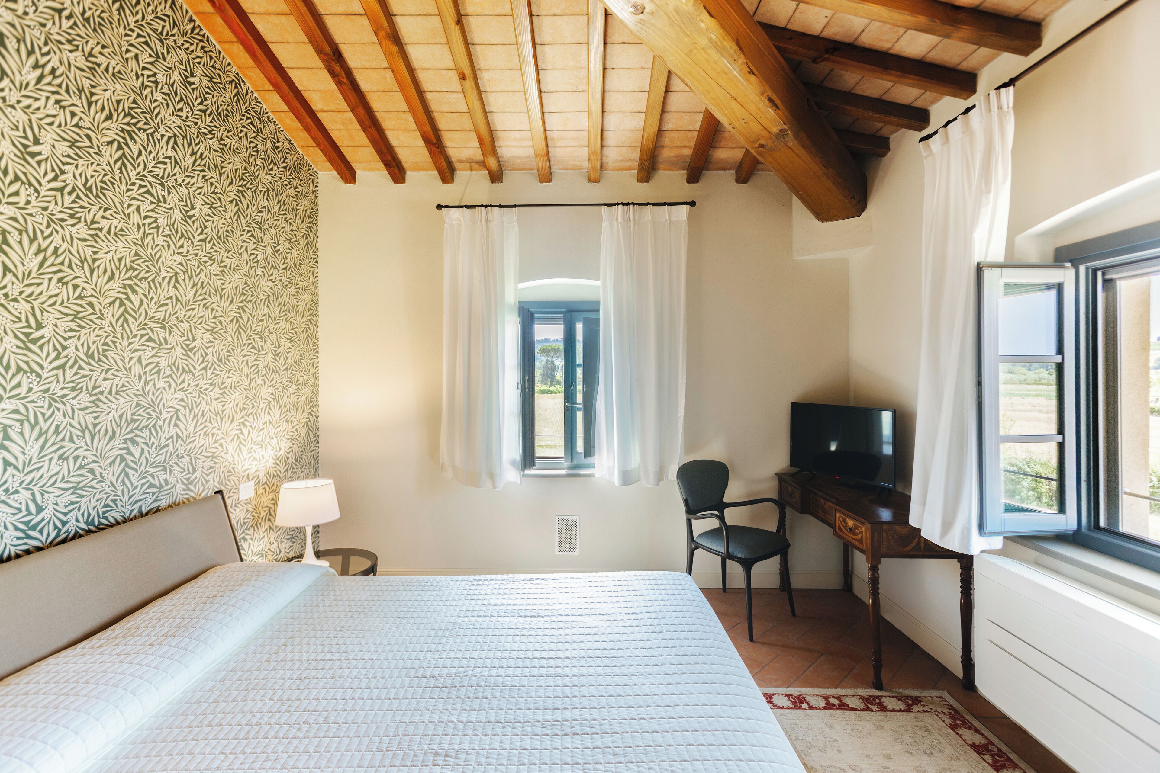 Un dormitorio con cama, escritorio, silla, televisor y ventana con cortinas.