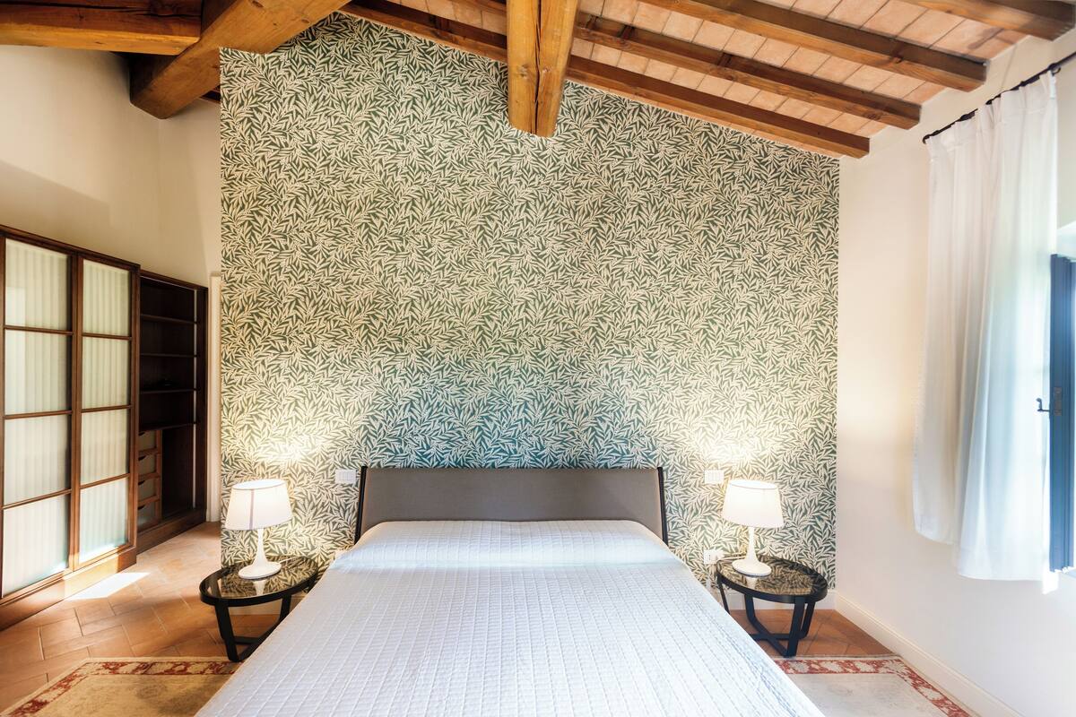 A bedroom with a patterned wallpaper, a bed with a white bedspread, two bedside tables with lamps, and a wooden ceiling beam.
