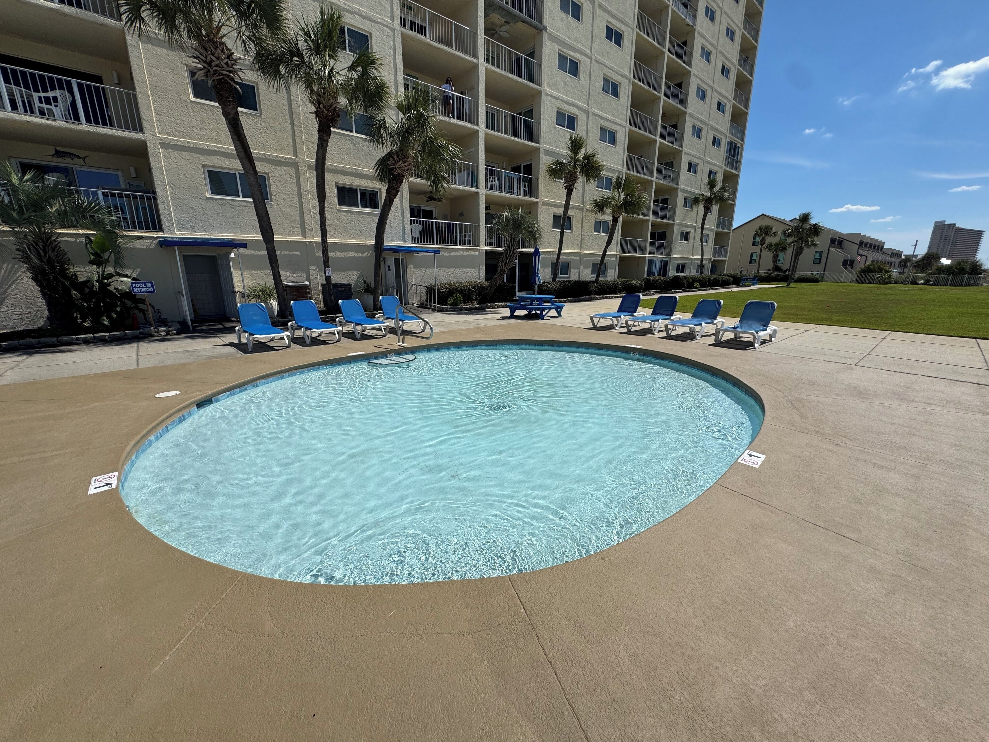 A swimming pool with blue lounge chairs, palm trees, and a multi-story building in the background.