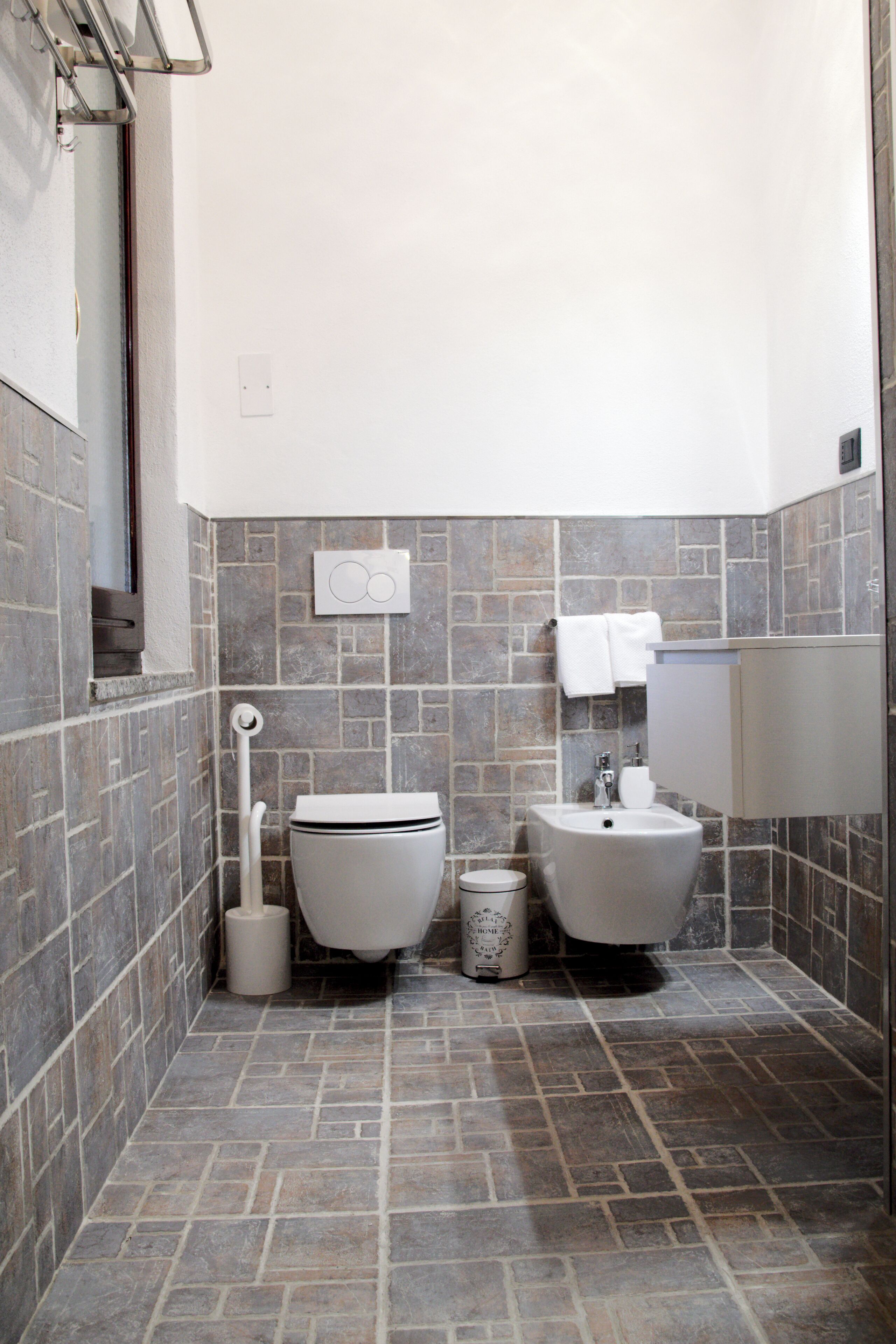 Un baño con inodoro, bidé y lavamanos, todo con accesorios blancos. El suelo y las paredes están cubiertos de baldosas grises que imitan la piedra.