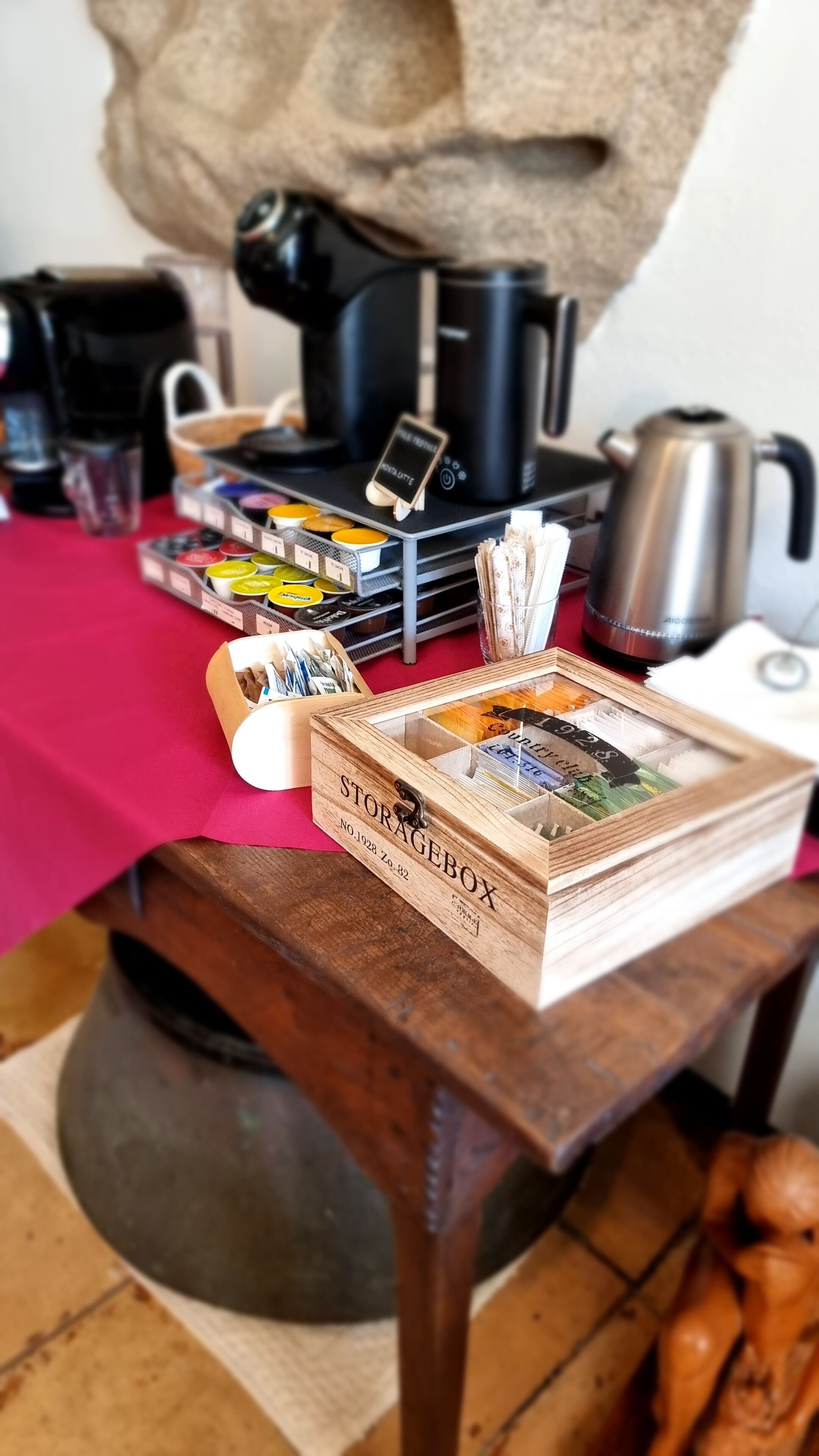 Une boîte en bois étiquetée « STORAGEROX », dotée de compartiments, une machine à café, une bouilloire et un téléphone intelligent, le tout posé sur une table recouverte d’un tissu rouge.