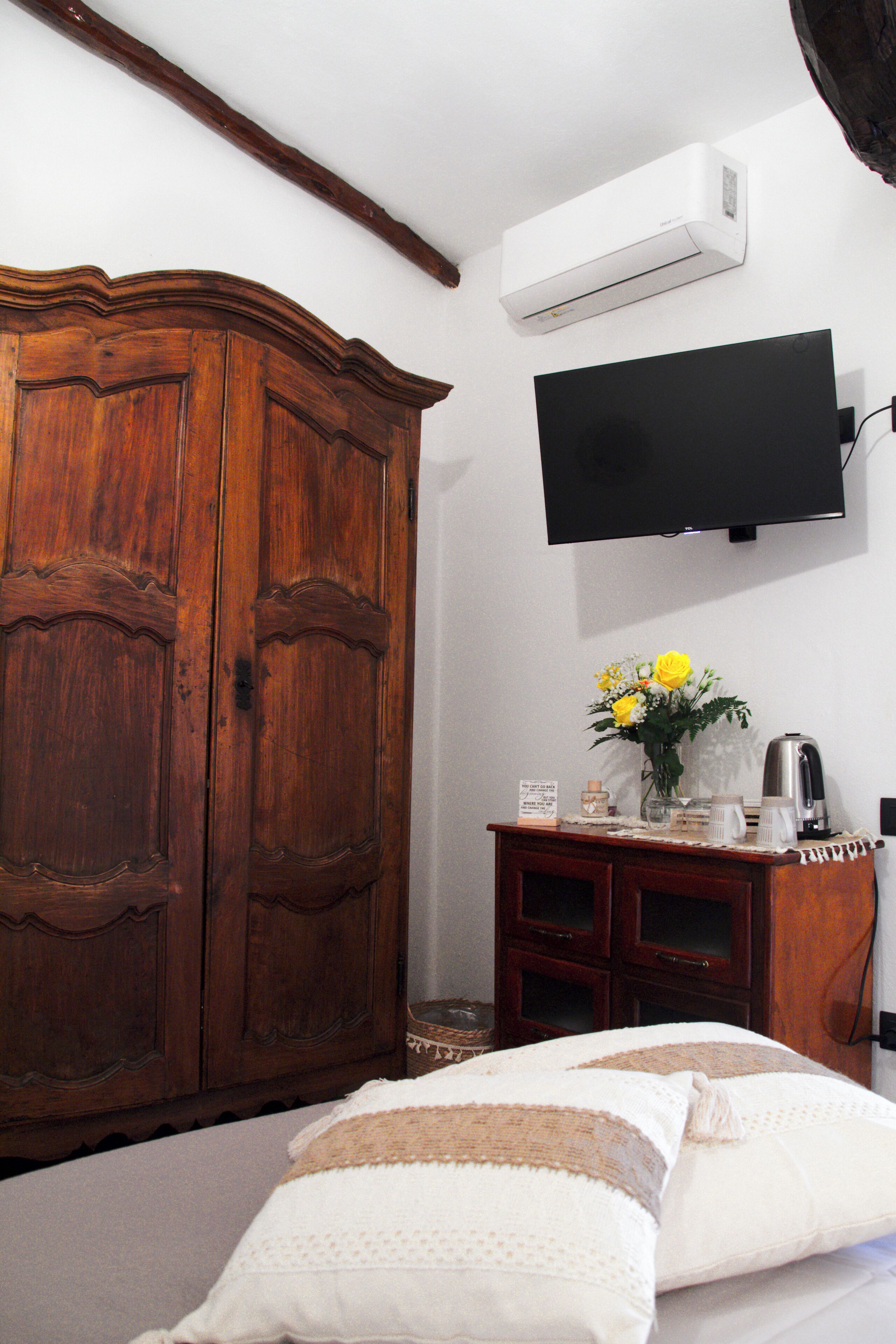 A bedroom with a wooden wardrobe, a flat-screen TV mounted on the wall, a bedside table with a coffee maker, and a bed with white bedding.