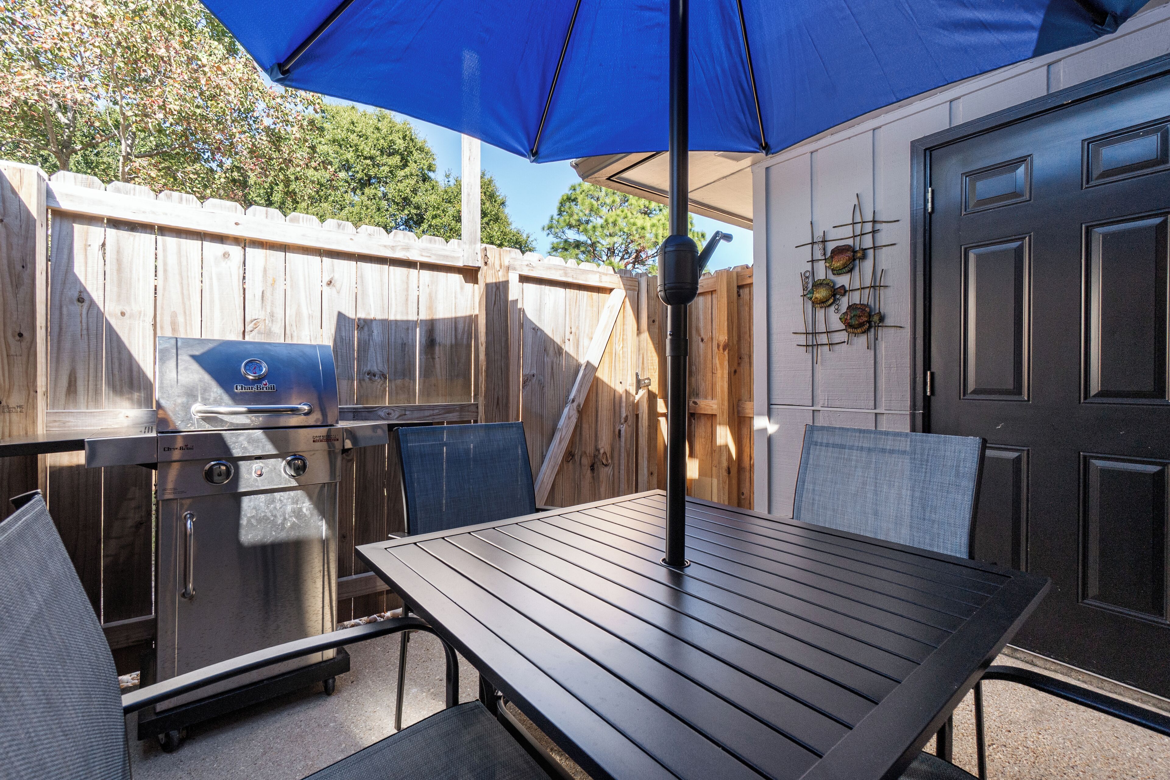An outdoor patio with a grill, a table, and chairs under a blue umbrella.