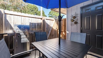 An outdoor patio with a grill, a table, and chairs under a blue umbrella.