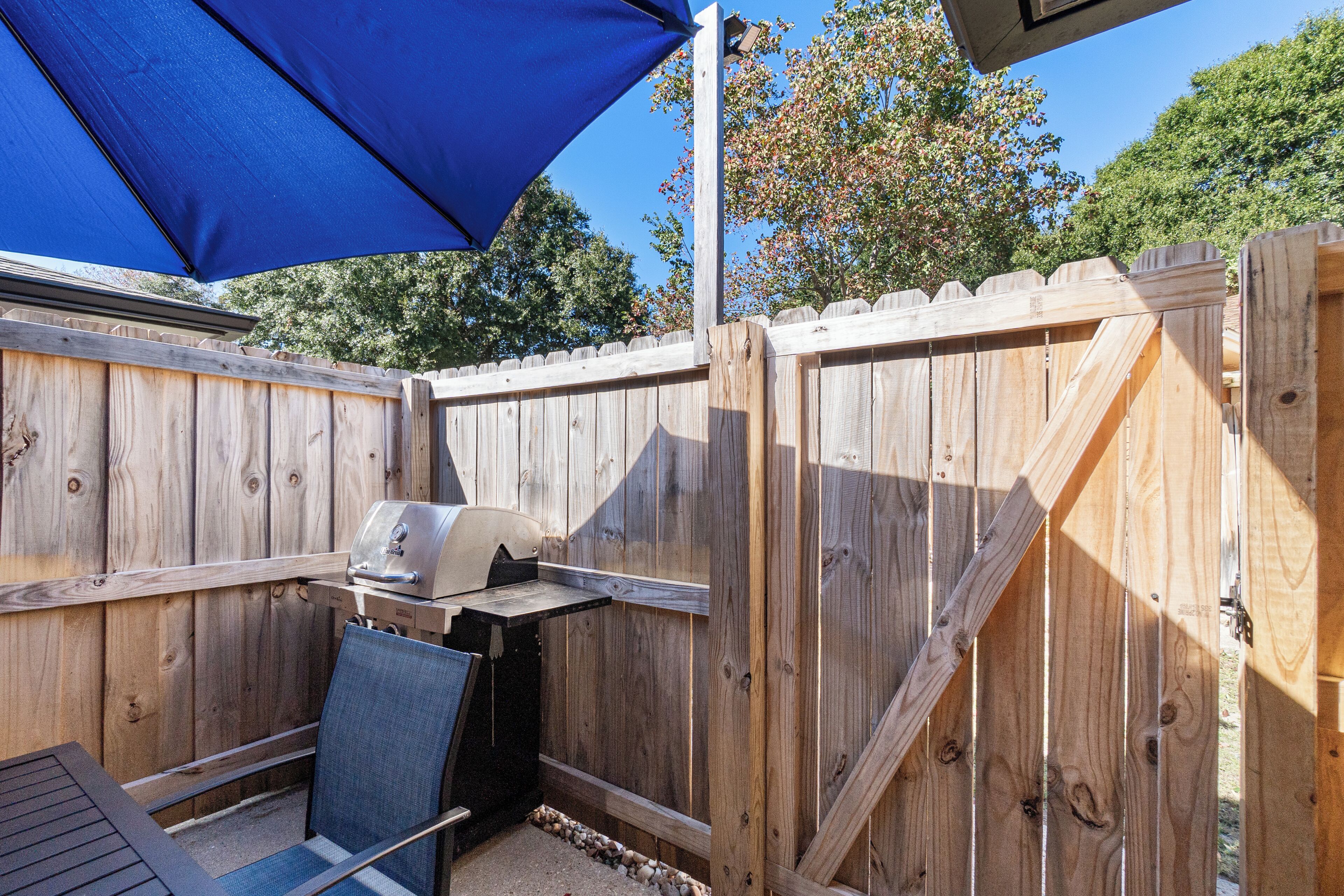 An outdoor patio with a wooden fence, a grill, and a blue umbrella providing shade.
