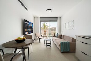 Una sala moderna con un sofá, una mesa de comedor con un tazón de fruta, un televisor montado en la pared y vistas al exterior a través de amplias ventanas.