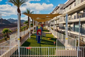 A children's play area with a slide and climbing structures, surrounded by a white fence and palm trees. - Eilat Bay Retreat • Sea Views by SeaNRent (Eilat)