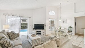 A spacious living room with a sofa, a coffee table, and a TV. There is a dining area with a table and chairs, and a kitchen area with a counter and chairs. - 4BR Lake Berkley Home, Pool & Game Room (Kissimmee)