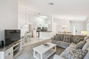A modern living room with a flat-screen TV, a sofa, a coffee table, and a kitchen in the background. - 4BR Lake Berkley Home, Pool & Game Room (Kissimmee)