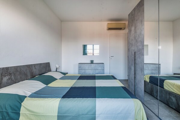 A modern bedroom with a large bed, a bedside table, a mirror, and an air conditioning unit on the wall.