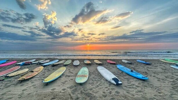 Una playa con varias tablas de surf alineadas en la arena, un atardecer al fondo y el océano.