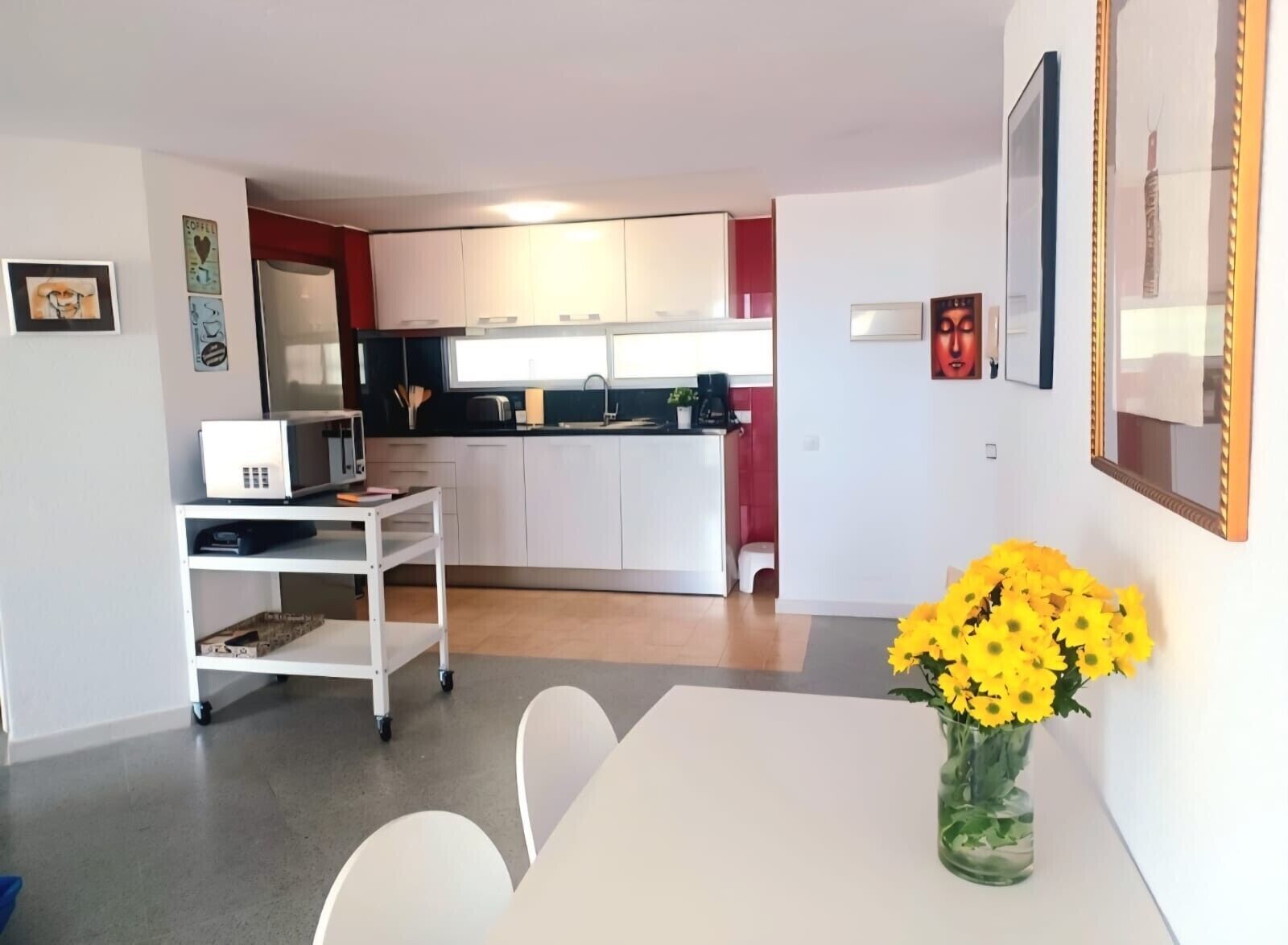 Une cuisine moderne avec des meubles blancs, un four à micro-ondes et une machine à café. On y trouve un coin repas avec une table et des chaises blanches, ainsi qu'un vase rempli de fleurs jaunes.