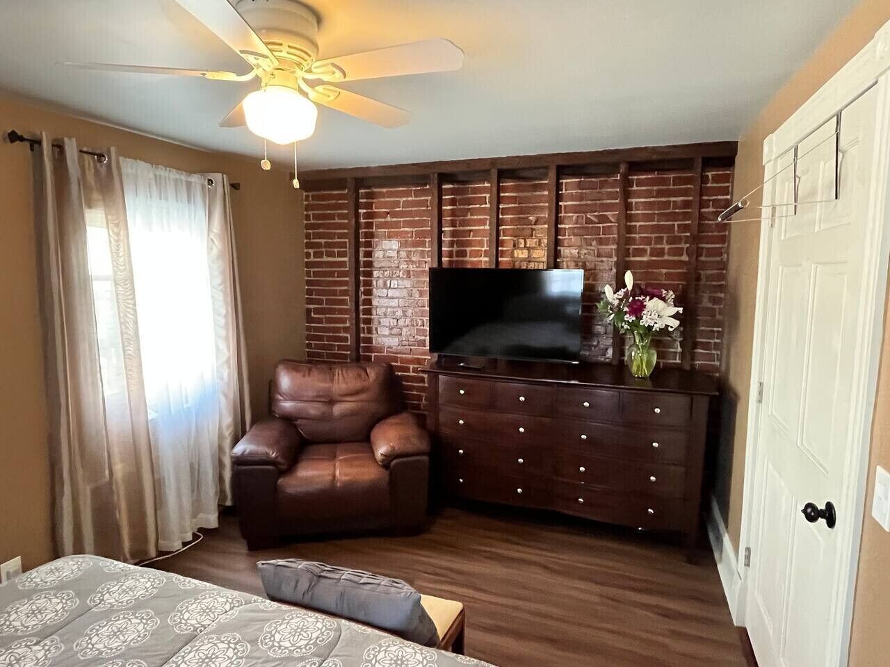 Une chambre avec un mur décoré d’un motif en briques, un canapé en cuir, un téléviseur posé sur un meuble en bois et un ventilateur de plafond.