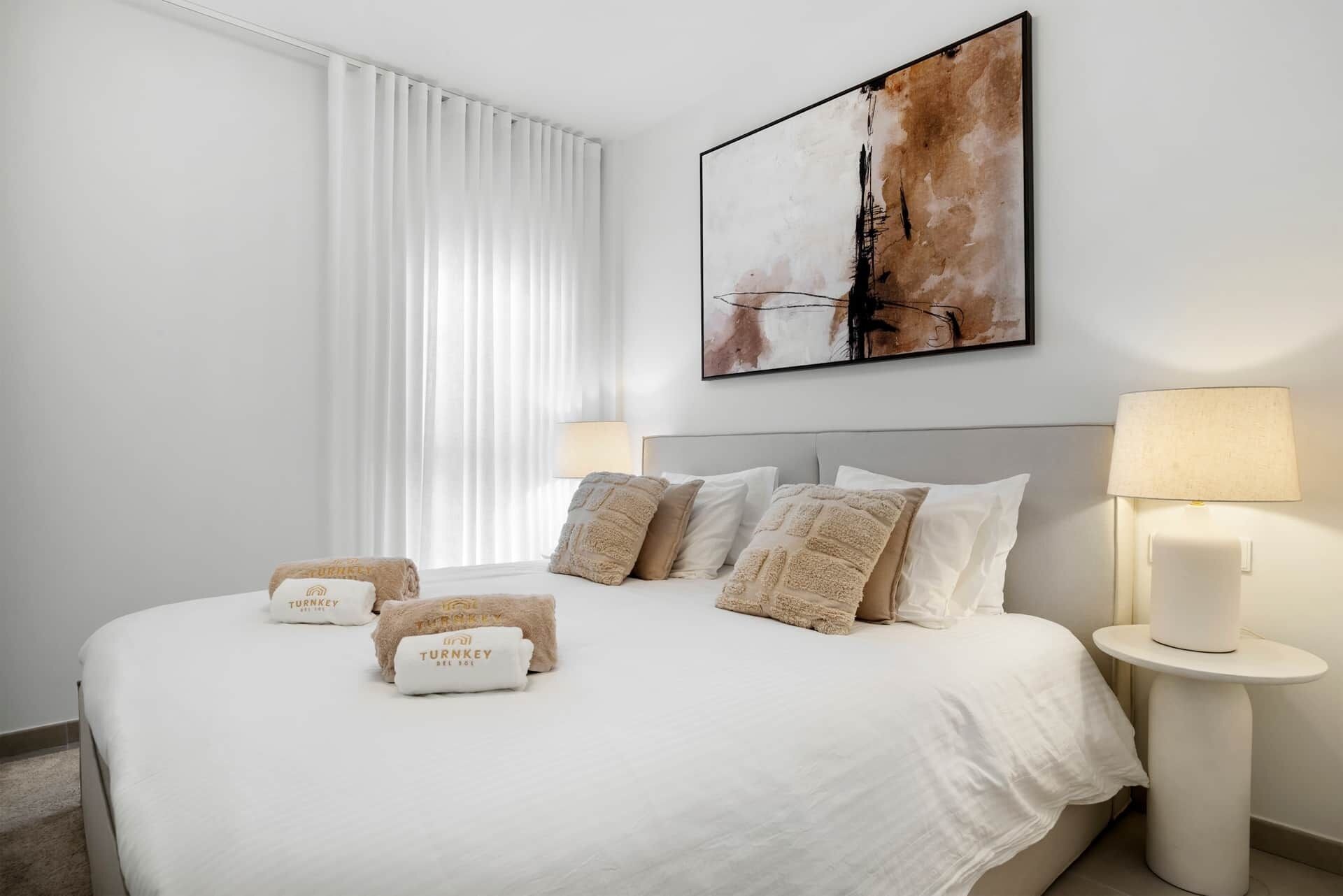 A hotel room with a large bed, white bedding, and a painting above it. There are two lamps on either side of the bed.