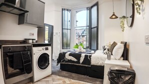 Ein kompaktes Hotelzimmer mit Waschmaschine, einem Bett mit schwarz-weiß gemustertem Überwurf, einem kleinen Nachttisch mit einer Vase und Blick ins Freie durch ein großes Fenster.