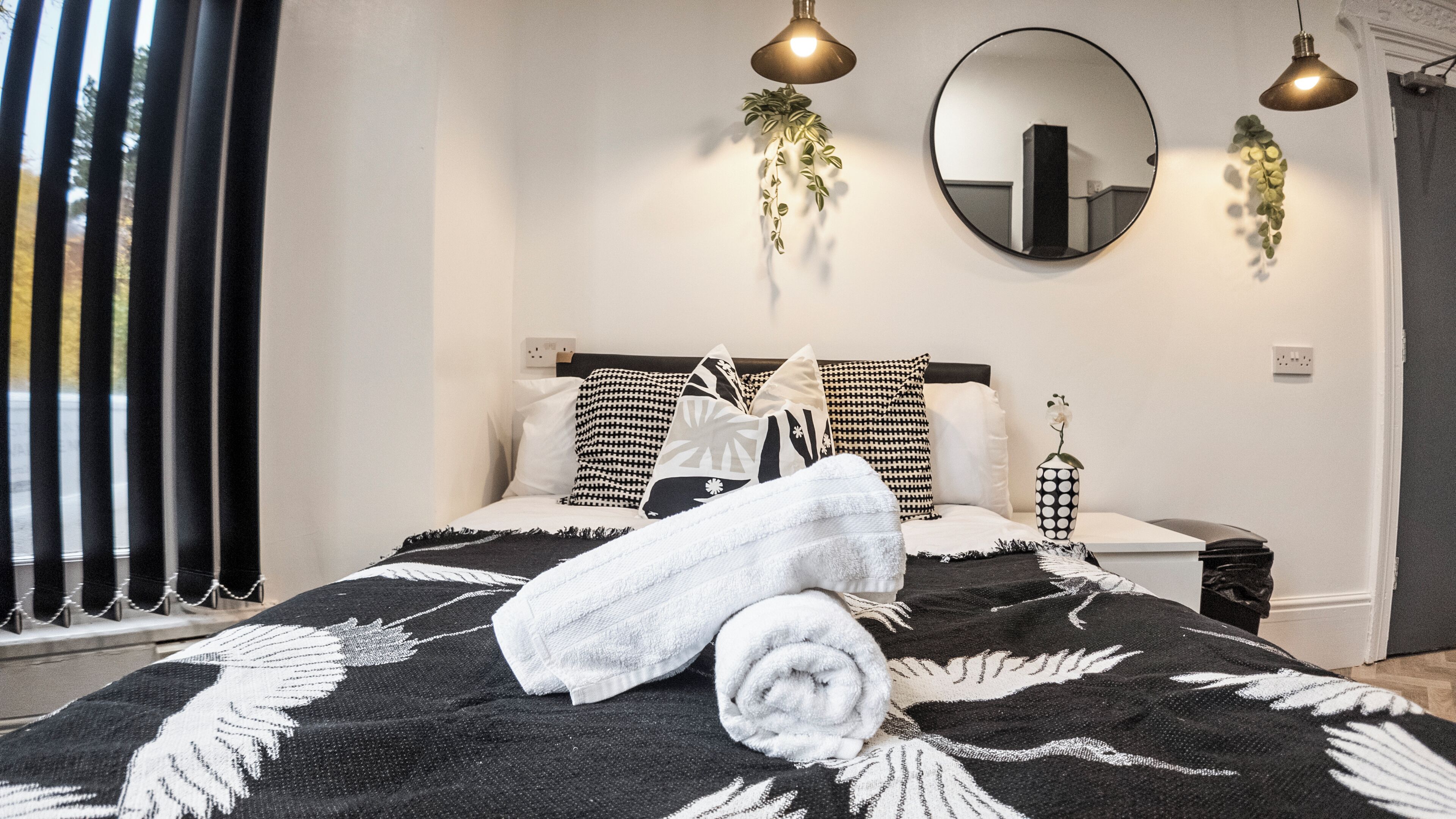 Ein ordentlich gemachtes Bett mit einer schwarz-weißen Decke, einem weißen Handtuch und mehreren Kissen in einem modernen Schlafzimmer mit einem großen Spiegel, hängenden Pflanzen und Pendelleuchten.