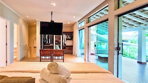 A spacious bedroom with a large bed, a wooden chair, and a TV. There is a closet with clothes and a bench outside the room.
