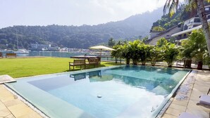 A rectangular infinity pool with a clear edge, surrounded by a pebble border. There are lounge chairs and a table with umbrellas near the pool. The background features a lush green hill and a body of water with boats.
