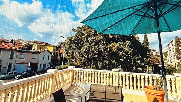 Ein Balkon mit Tisch und Stühlen, einem grünen Sonnenschirm und Blick auf Häuser und Bäume.