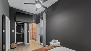 A bedroom with a bed, a ceiling fan, a wooden floor, and a hallway leading to a bathroom. - Modern Soulard • WiFi • Laundry • Queen • Menard (St. Louis)