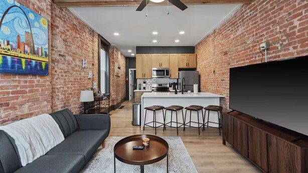 A modern living room with a gray sofa, a coffee table, and a TV. The room has exposed brick walls and a ceiling fan. - Modern Soulard • WiFi • Laundry • Queen • Menard (St. Louis)