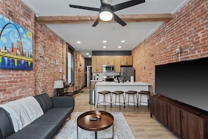 A modern living room with a gray sofa, a coffee table, and a TV. The room has exposed brick walls and a ceiling fan. - Modern Soulard • WiFi • Laundry • Queen • Menard (St. Louis)