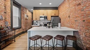 A modern kitchen with a brick wall, wooden cabinets, stainless steel appliances, and a bar area with stools. - Modern Soulard • WiFi • Laundry • Queen • Menard (St. Louis)