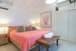 Ein ordentlich eingerichtetes Schlafzimmer mit einem hölzernen Bett, gemusterten Kissen, einer rosa Decke, einem Nachttisch mit einem Handtuch und einem Wandbild.