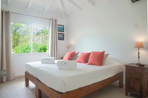 A bedroom with a large bed, two bedside lamps, a window with curtains, and a view of greenery outside. - Villa Les Lataniers (W582+QP6 Gustavia, St Barthélemy)