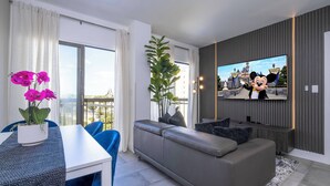 Ein modernes Wohnzimmer mit einem grauen Sofa, einem Esstisch mit blauen Stühlen, einem an der Wand montierten Fernseher und einem Balkon mit Vorhängen.