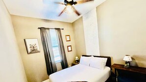 Camera d'albergo con un letto, un comodino, una lampada, una ventola a soffitto e una finestra con tende.