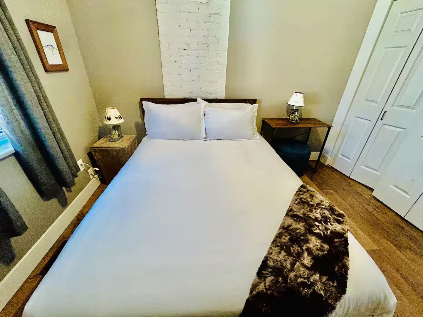 A neatly made bed with white linens, a wooden headboard, a small bedside table with a lamp, and a large window with curtains.